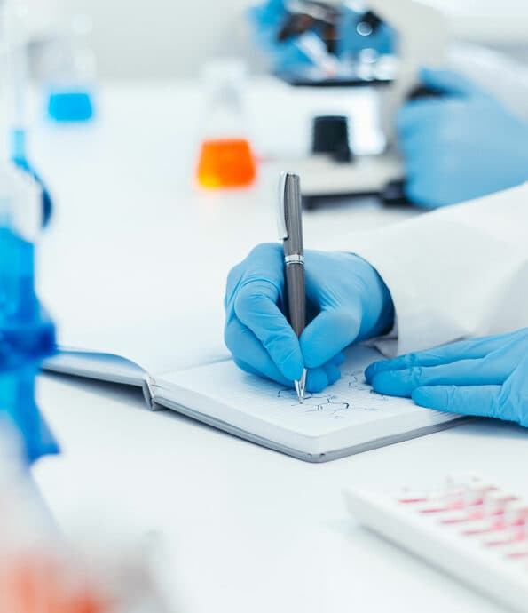Gloved hands drawing a chemical structure in a lab notebook beside a microscope and flasks